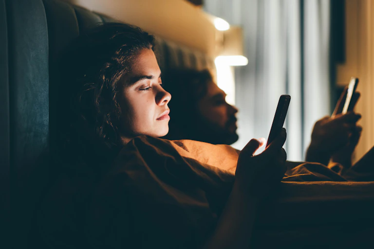 A man and woman lie in bed staring at smartphones