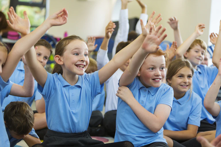 Happy children in blue t-shirts raise their hands in a classroom.