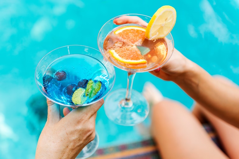 Hands holding colourful mocktails above feet in a pool.
