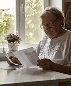 Elderly woman sitting at table in kitchen at home holds domestic bills
