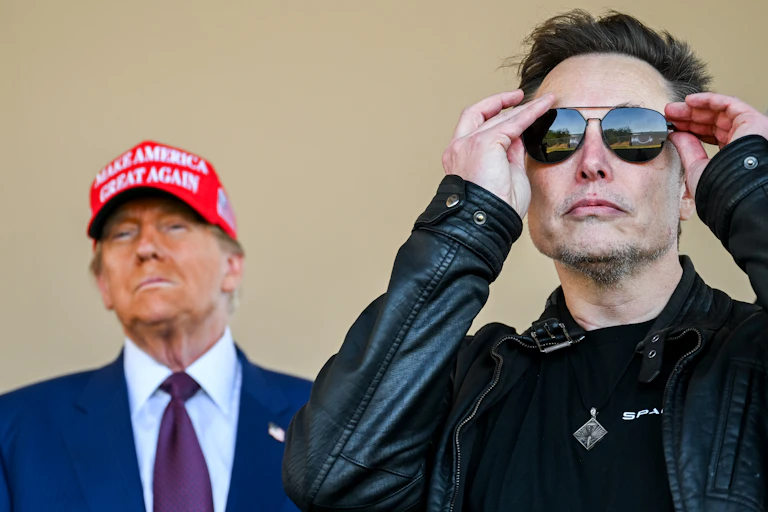 Two men, one in the background wearing a 'Make America Great Again' hat, and one in the foreground putting on sunglasses.