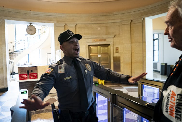 A man in a uniform and a badge holds out his arms, palms outstretched.