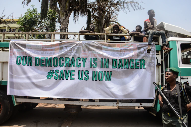 Men on the back of a mini truck with a large white banner saying Our democracy is in danger, save us now