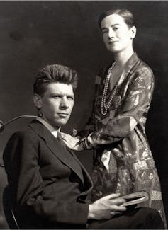 A young woman poses standing next to a young man with spiky hair who's seated.