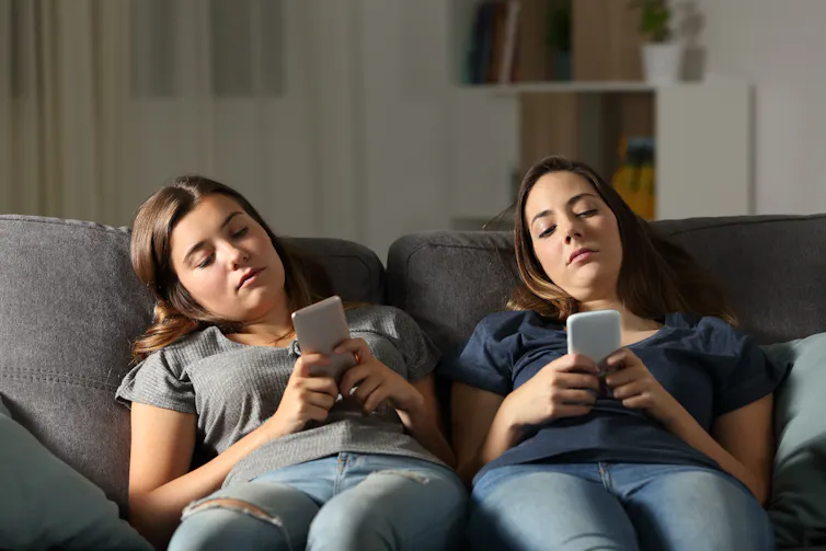 two bored young women sit on a couch using smartphones