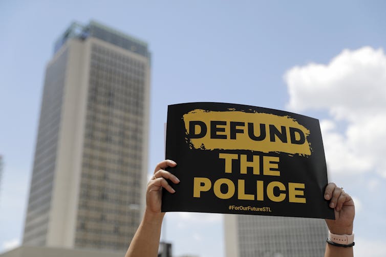 A person holds up a defund the police sign with a tall building in the background.