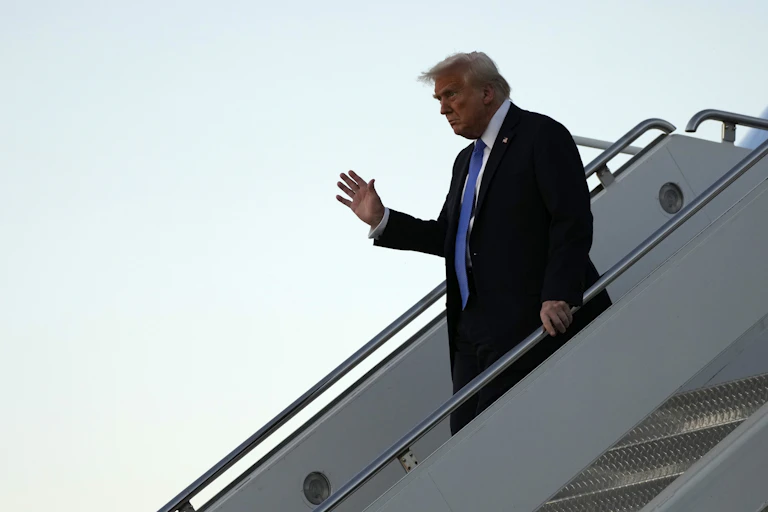 A hulking man with prominent fluffy white-blond hair waves as he walks down a set of stairs from an aircraft.