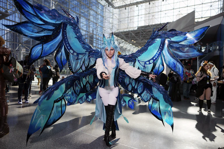 A woman in a white outfit with huge blue wings attached to her poses in a large convention atrium.
