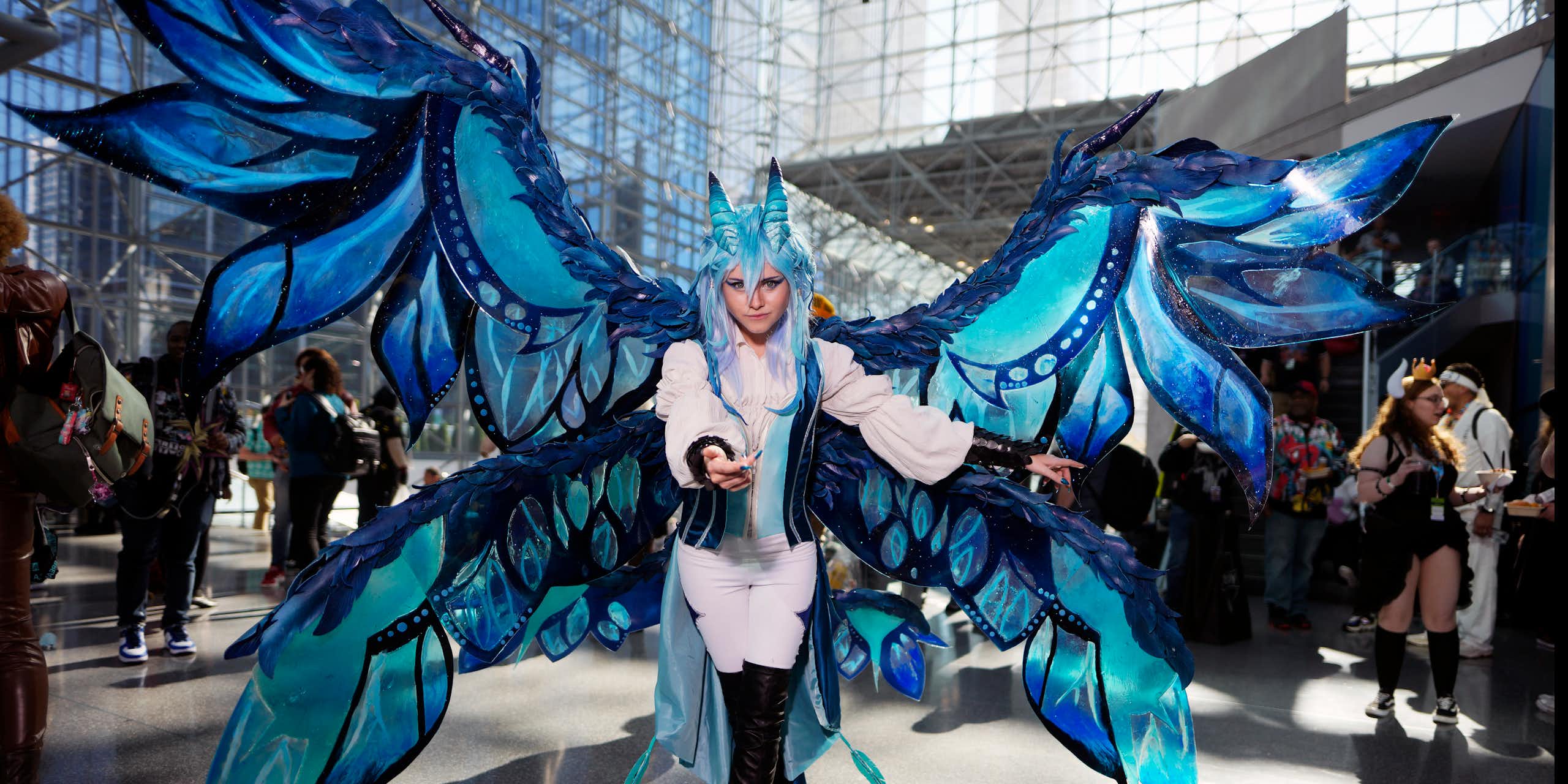A woman in a white outfit with huge blue wings attached to her poses in a large convention atrium.