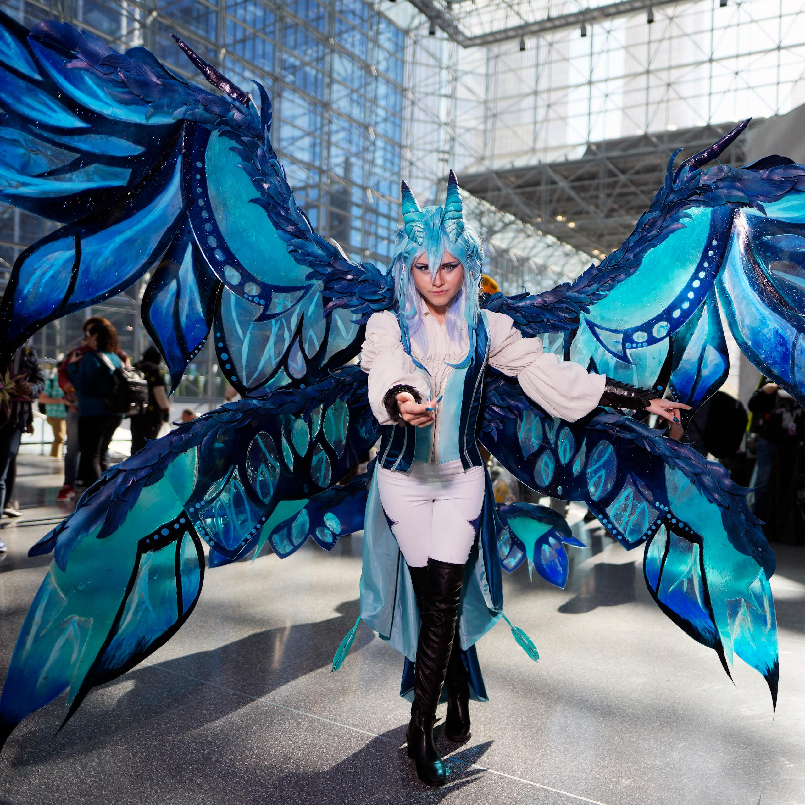 A woman in a white outfit with huge blue wings attached to her poses in a large convention atrium.