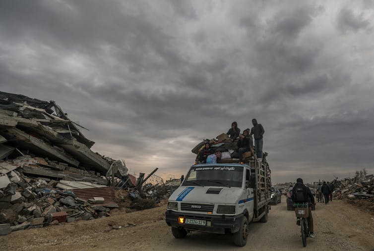 A pickup truck loaded with people drives along a dirt road among destroyed buildings.