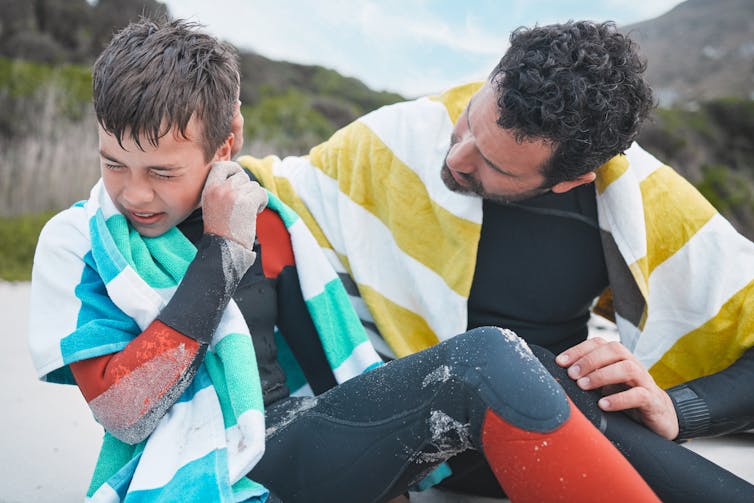 A young boy tries to get water out of his ear while his father tries to help.