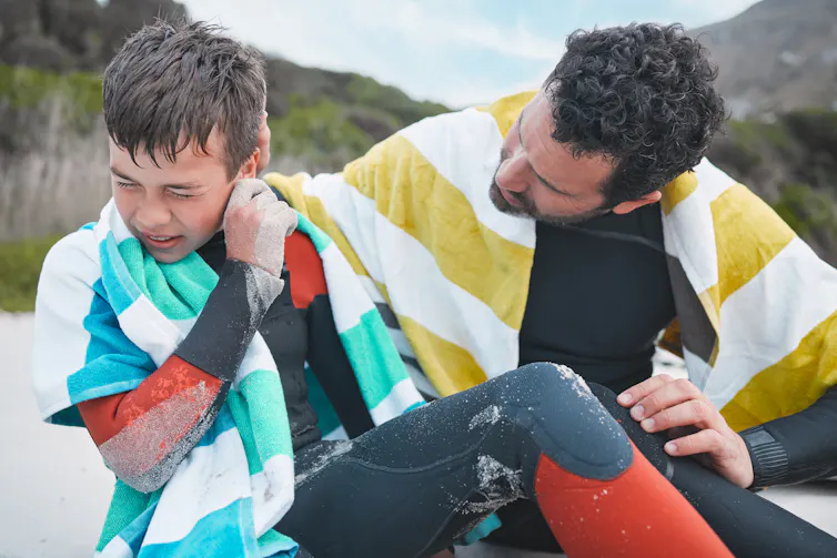 A young boy tries to get water out of his ear while his father tries to help.