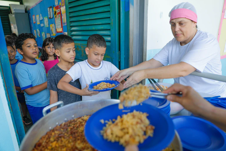 Children wait in line to be served food by a worker wearing a pink hairnet.