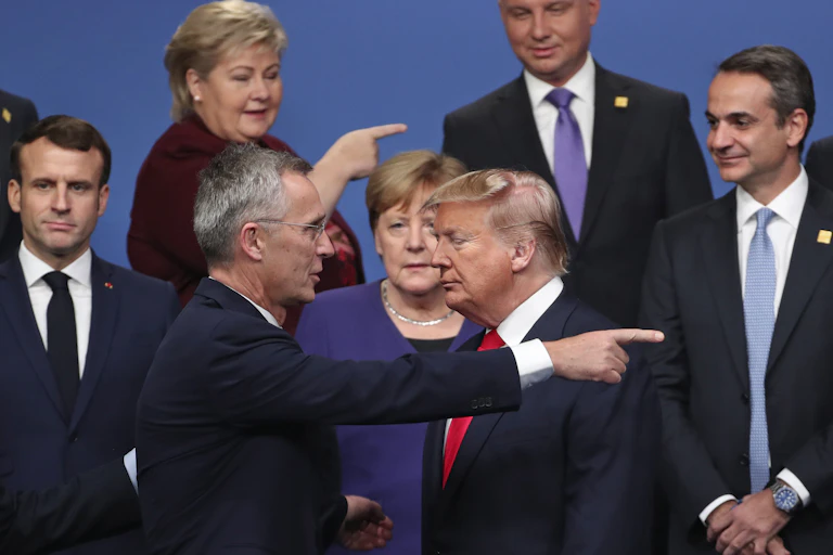 A man with fluffy, swirling orange-ish hair stands with his eyes shut while two people close to him point in the opposite direction.