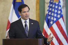 A dark-haired man speaks from behind a lectern with an American flag behind him.