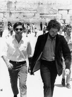 A black and white photo of two men in dress pants and button-up shirts walking through a large outdoor plaza, with a stone wall in the background.