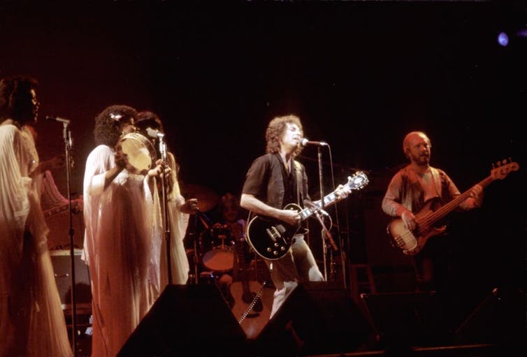 Two men play guitar on stage next to a group of women singers in white dresses.