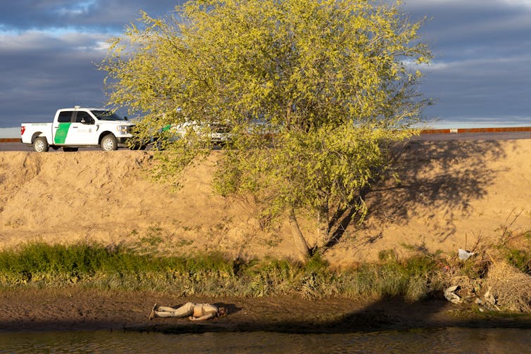 A white-and-green pickup truck is at the top of a river bank. Near the river is a body lying face down.