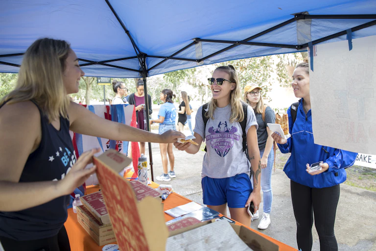 A young woman behind a table under a tent hands pizza to another young woman wearing a backpack, while other students mill in the background