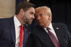 An older man with blondish-grey fluffy leans into speak to a younger bearded dark-haired man.