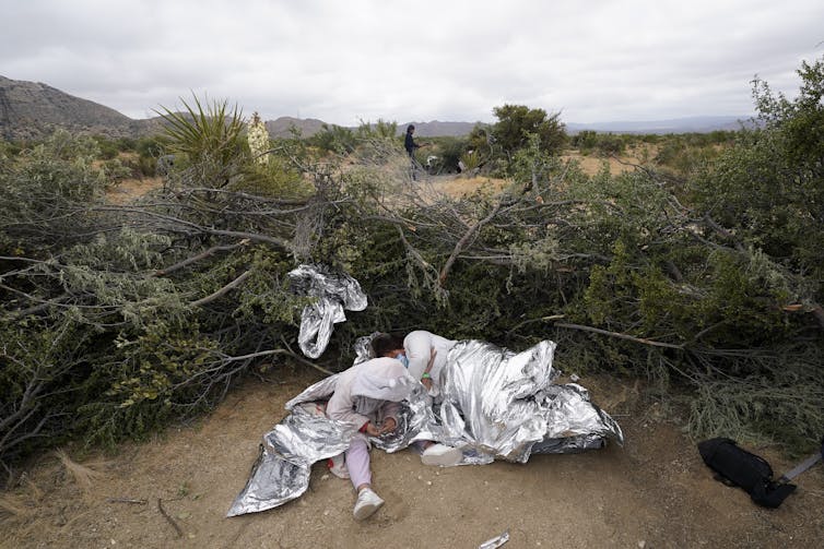 A woman wrapped in a foil blanket sleeps on the floor next to a child, surrounded by dry shrubs.