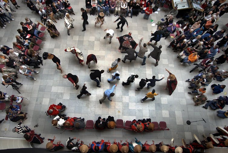 A bird's-eye view of around a dozen people dancing in an atrium as rows of seated people around them watch.
