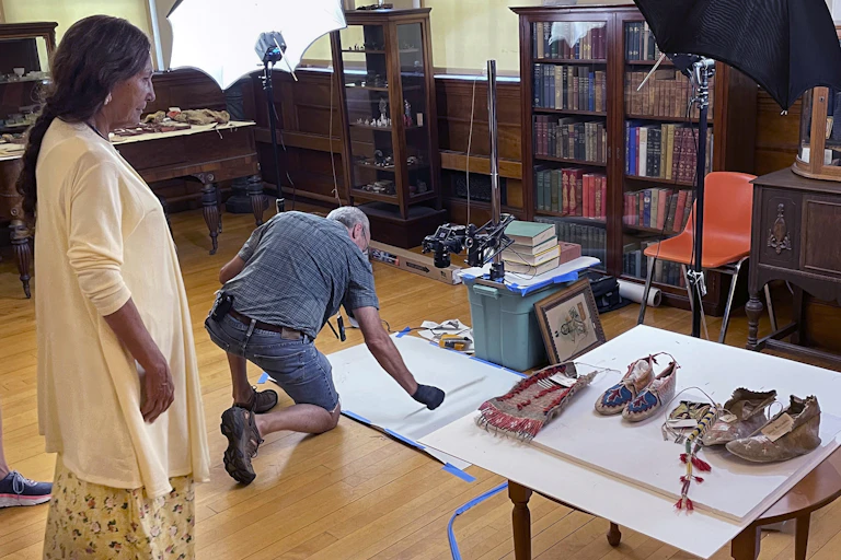 A woman stands in a room with wooden trim and furniture as a man in jean shorts crouches over a mat on the ground.