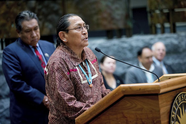 A man in a red shirt with glasses and a blue beaded necklace speaks into a mic at a wooden podium.
