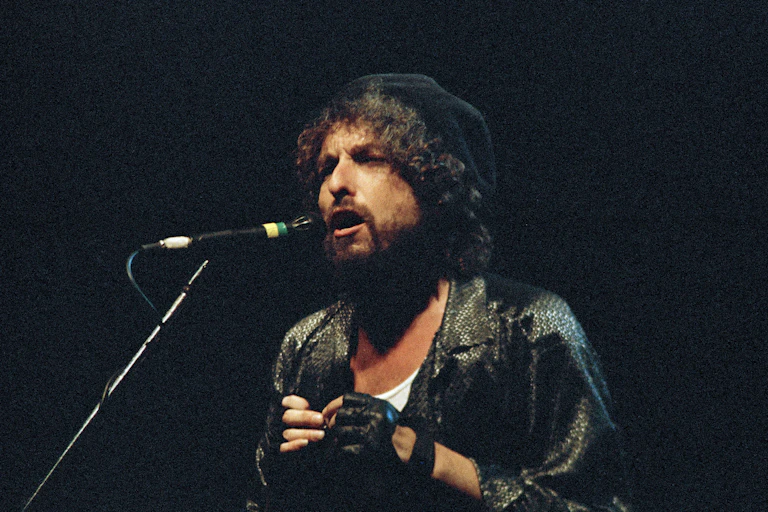 A man with curly hair wearing a shiny dark jacket and gloves sings into a microphone.