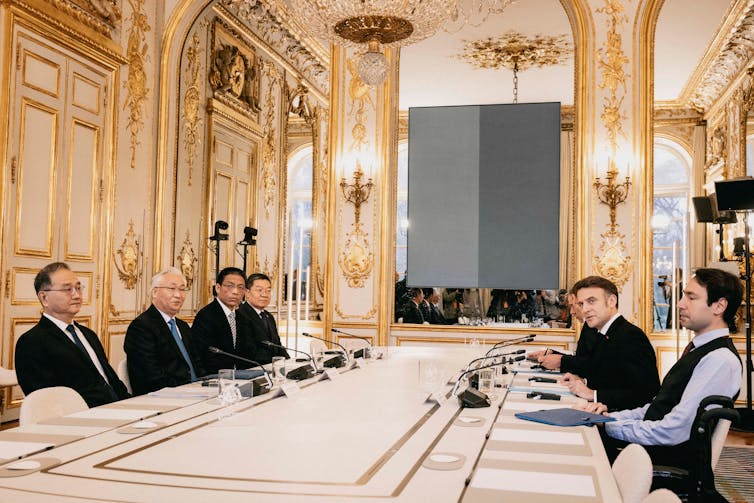 O vice-primeiro-ministro da China, Zhang Guoqing, e o presidente da França, Emmanuel Macron, com conselheiros, sentam-se um em frente ao outro em uma grande sala espelhada e dourada