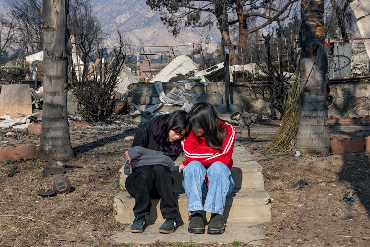 Two young women sit on the steps of a burned home.