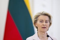 A woman with short blonde hair stands in front of a red, green and yellow flag.