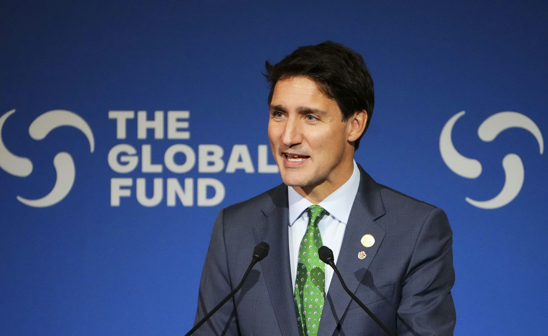 A man in a dark suit and a green tie speaking in front of a sign reading The Global Fund