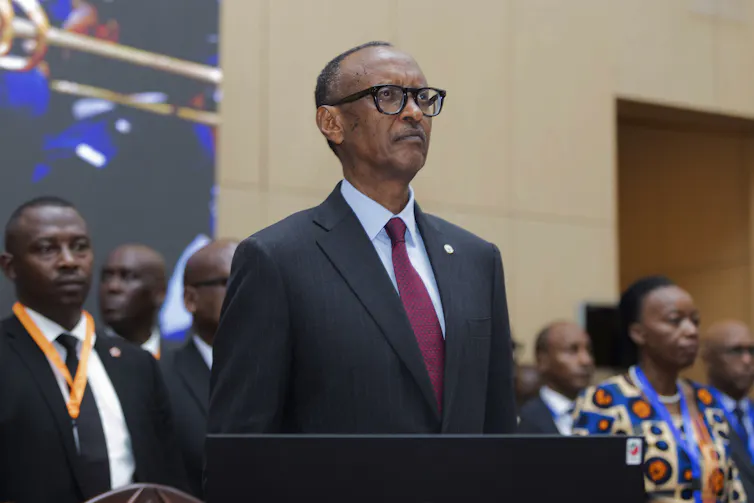 A man wearing glasses, a dark suit and red tie standing in a room with other people in the background