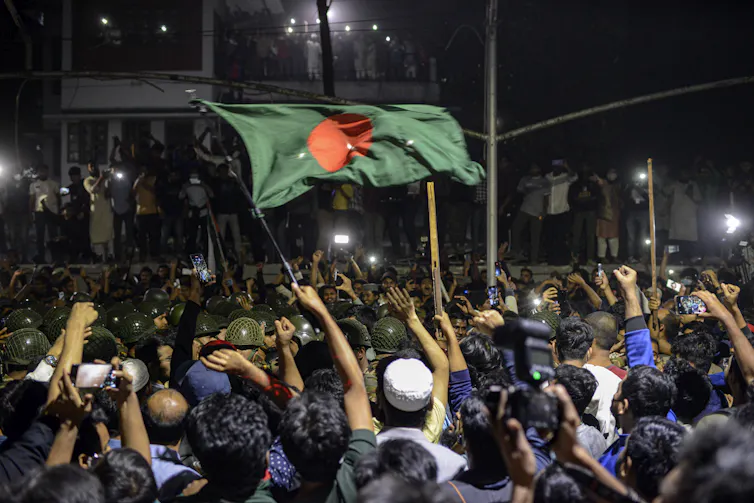 Protestors waving a green flag with a red circle in the middle, clashing with police outside a vandalised home.