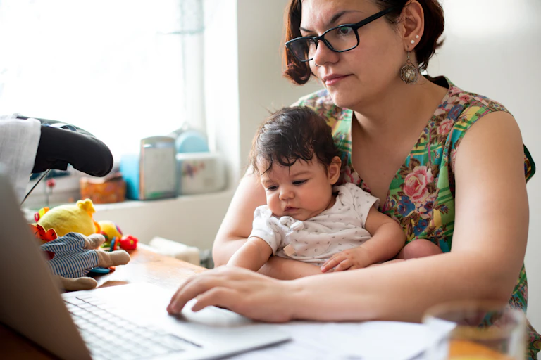 A woman holding a baby looks at her laptop.