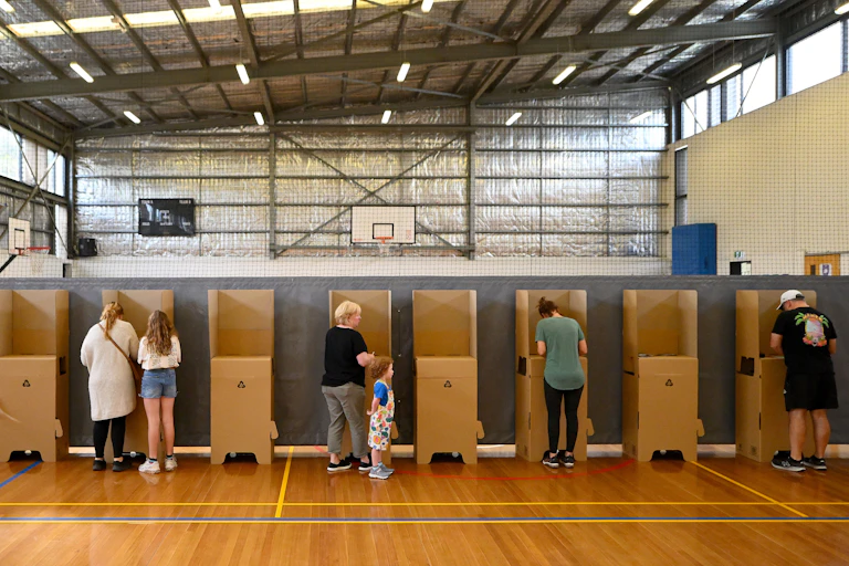 Adults vote in a line in a school gym. A young girl watches her mother vote in a booth.