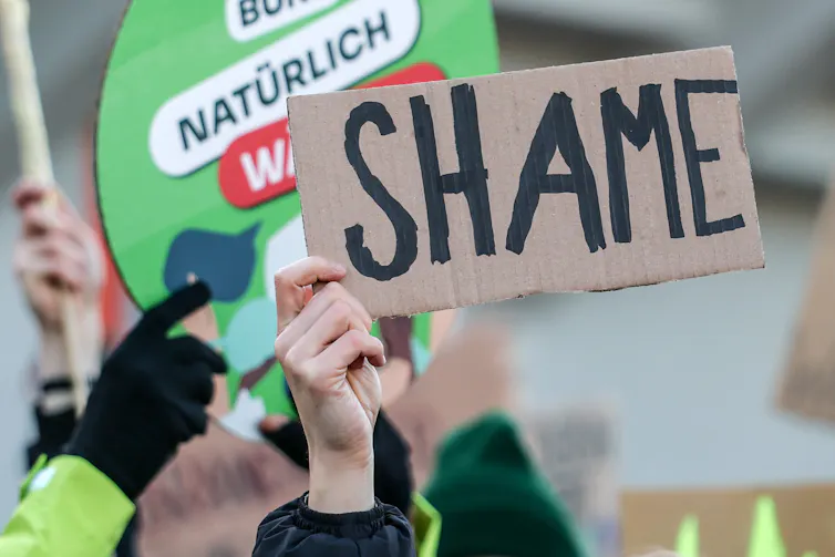 A hand holding up a cardboard sign that reads 'shame'
