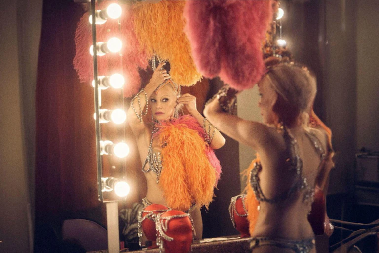 Pamela Anderson dressed in showgirl feathers, putting on an earring in the mirror.