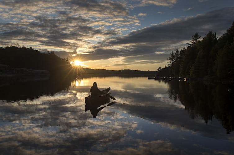 A person in a canoe as the sun sets on a lake with forest on either side.