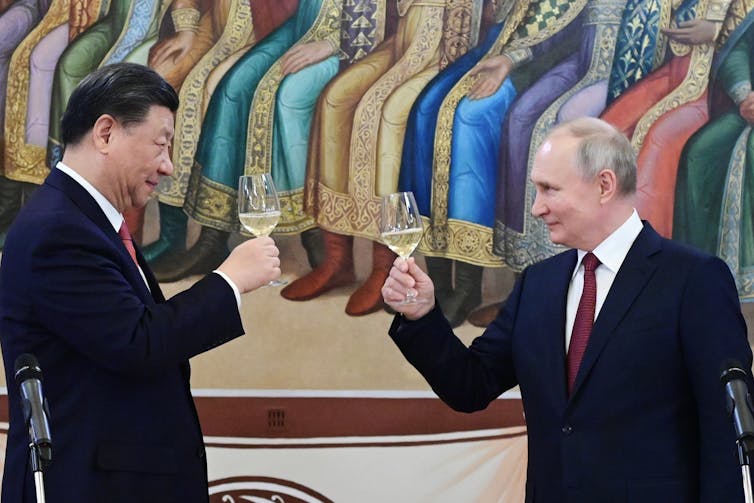 A tall Asian man and a balding shorter white man hold up glasses of champagne while smiling at each other.