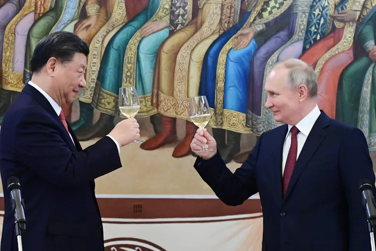 A tall Asian man and a balding shorter white man hold up glasses of champagne while smiling at each other.