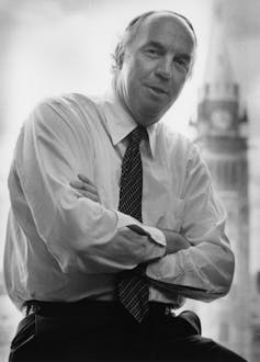 A black-and-white photo of a man with thinning hair and with his arms crossed, the Peace Tower behind him.