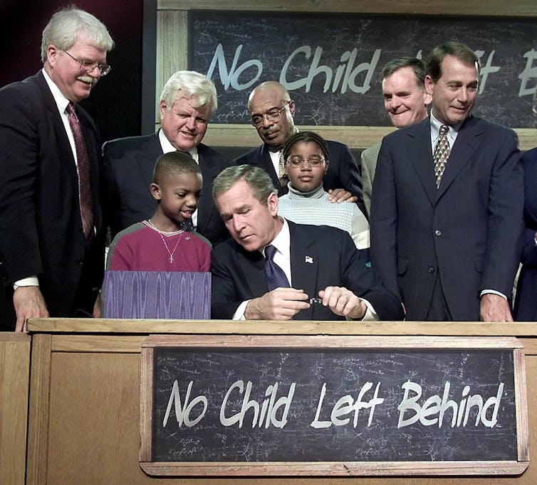 President George W. Bush sits at a desk surrounded by students and politicians.