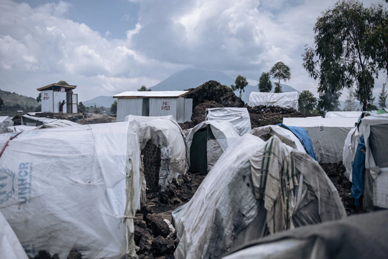 Small structures covered with white canvas written unhcr on muddy ground in the foreground, with a mountain in the background