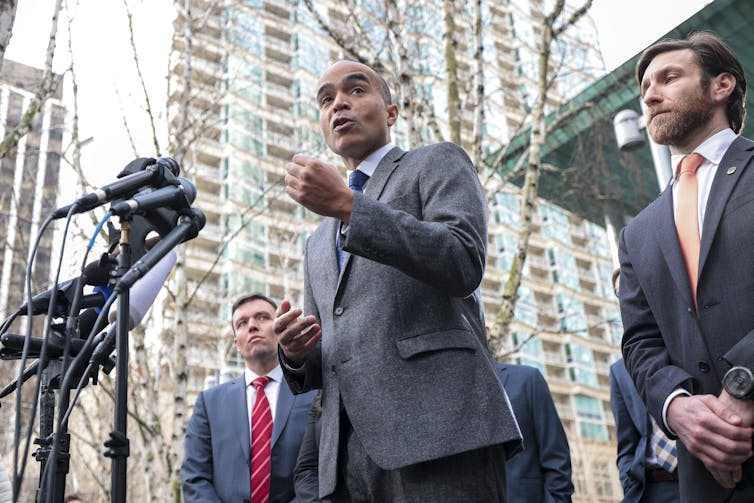 A man stands in front of several microphones outside of a large building and appears to speak into them and gesture with his hand.