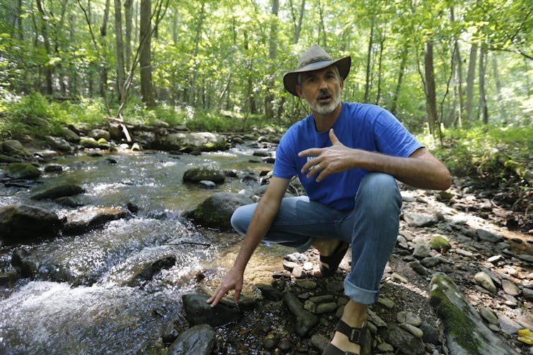 Un homme vêtu de jeans et d’un chapeau de cowboy agenouillé sur un terrain à côté d’un ruisseau