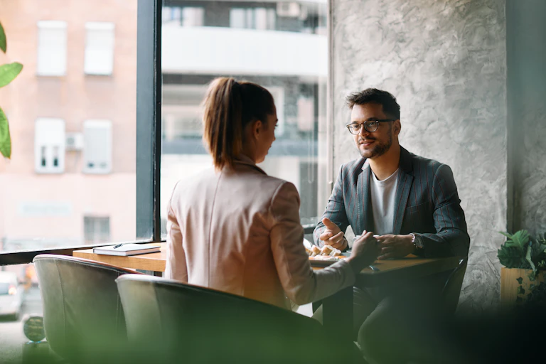 A man and a woman have a conversation while seated at a table near a window