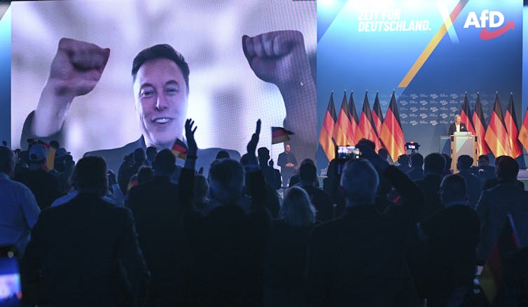 Man with raised fists on a screen at a large rally with lots of German flags.